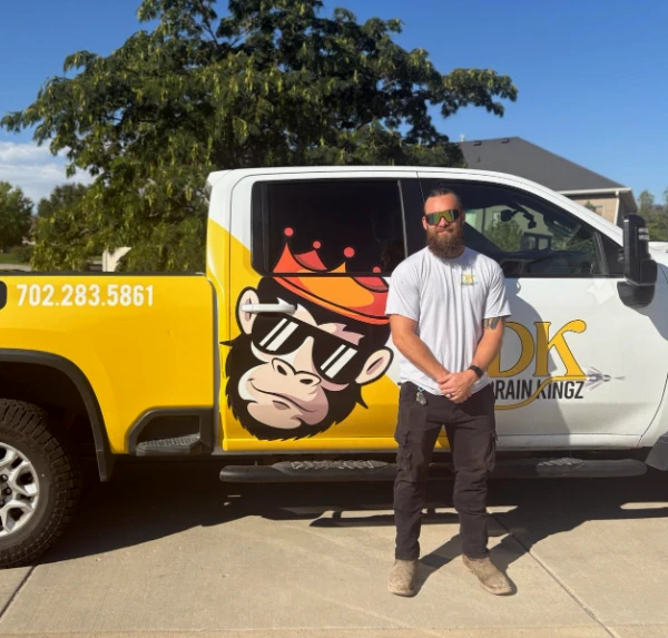 Bearded man in a white t-shirt and sunglasses stands in front of a yellow‑and‑white branded training truck featuring a monkey logo with sunglasses on the door panel.