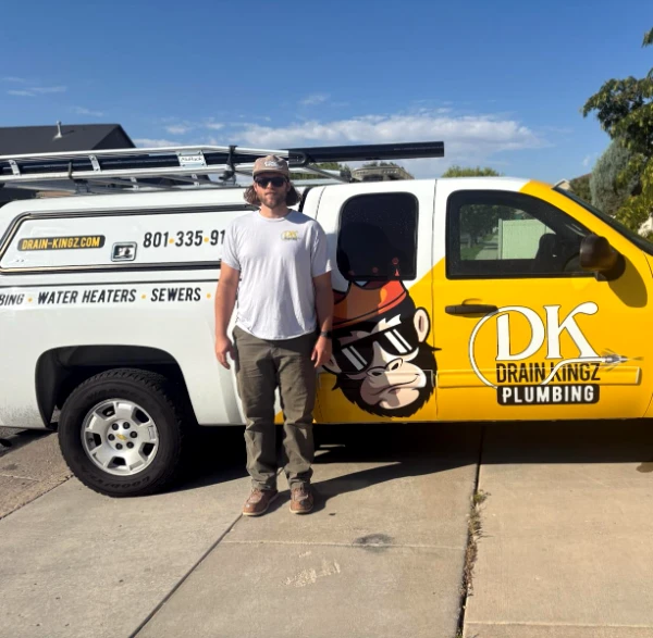 Man standing beside a white and yellow DK Drain Kingz Plumbing van with a monkey logo on a sunny driveway.