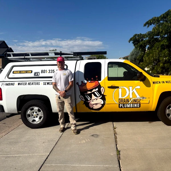 Plumber in gray shirt and tan pants standing beside a yellow and white DK Drain Kingz Plumbing truck with a cartoon dog logo on a sunny day.