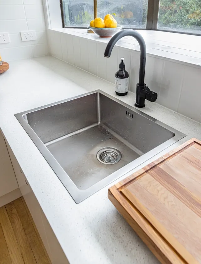 Stainless steel kitchen sink with a tall curved black faucet and a soap dispenser beside it on a light speckled countertop by a window with lemons on the sill.